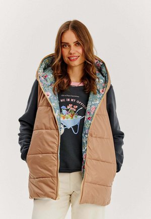 Beżowa kamizelka z ociepleniem i kwiatowym wzorem podszewki, z kapturem i zapięciem na zamek. Noszona na ciemny hoodie i kremowe spodnie.
