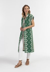 Abito lungo senza maniche di colore verde con un motivo a diamanti, indossato sopra una maglietta bianca e pantaloni bianchi, abbinato a sandali color beige.
