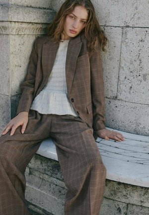 Brown checkered suit with a tailored jacket and wide-leg trousers, paired with a light textured sweater, set against a stone backdrop.