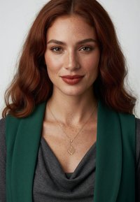 Halskæde med vedhæng formet som et guldløv på en person med kastanjebrunt hår, iført en grøn slå-om og en grå top med høj krave. Fregner er synlige.