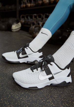 Baskets Nike blanches et noires portées avec des chaussettes Nike blanches et des leggings bleus sur le sol de la salle de sport avec des kettlebells en arrière-plan.