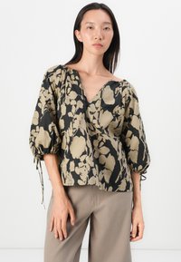 Haut à motif floral noir et beige avec des manches bouffantes, décolleté en V profond et accents à nouer sur les manches. La texture du tissu semble lisse.