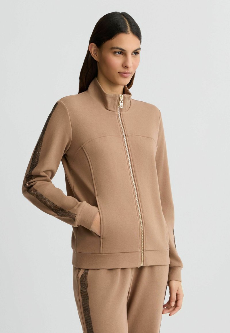 Tan zip-up jack met een zachte stof, hoog kraag, zijzakken en gestructureerde gestreepte accenten op de mouwen. Soepele, casual uitstraling.