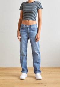 Randig cropped T-shirt i blått och grönt, högmidjade ljusblå denimjeans och vita sneakers, med en bakgrund av trägolv.