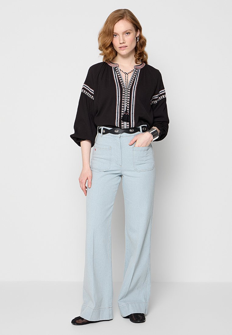 Schwarze Bluse mit Stickerei, V-Ausschnitt und lockeren Ärmeln, kombiniert mit hellblauen, weiten Hosen mit Fronttaschen und einem Gürtel.