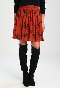 Jupe orange rouille avec un design plissé et un motif floral noir. Dispose d'une ceinture élastique. Associée à des bottes noires au-dessus du genou.