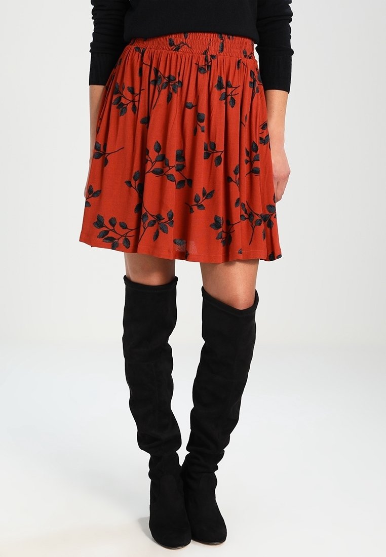 Jupe orange rouille avec un design plissé et un motif floral noir. Dispose d'une ceinture élastique. Associée à des bottes noires au-dessus du genou.
