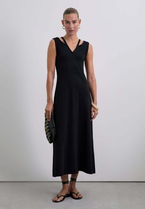 Zwarte mouwloze jurk met een V-hals, getailleerde lijfje, vloeiende rok en zijkanten met banden. Model houdt een gestreepte clutch vast en draagt platte sandalen.
