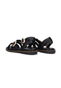 Zwarte sandalen van gestructureerd materiaal hebben twee grote goudkleurige ringen, een enkelband en een zwart studded zool.