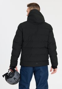 Schwarze Pufferjacke mit Kapuze, ausgestattet mit einem gesteppten Design und elastischen Bündchen. Eine Person hält einen grauen Skihelm in der rechten Hand.