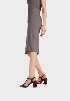 Donna che indossa un abito midi grigio aderente e sandali neri con tacco a blocco e punta aperta con cinturini alla caviglia, in piedi su uno sfondo semplice.