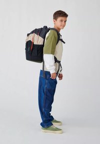 Sac à dos noir et beige avec des accents rouges, design rembourré, bretelles ajustables et forme rectangulaire. Porté par une personne en tenue décontractée.