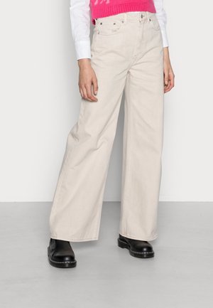 Persoon die beige wijd uitlopende broek, zwarte laarzen, wit shirt met lange mouwen en roze mouwloos vest draagt, staande binnen op een grijze vloer.