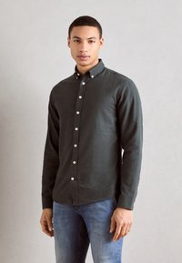 Dark green button-up shirt made of textured fabric, featuring a collar, straight cut, and white buttons. Paired with blue jeans.