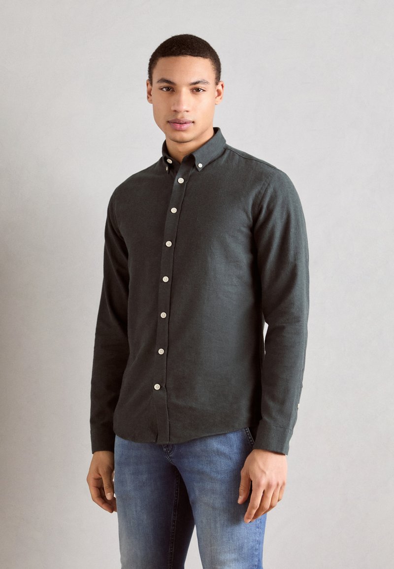 Dark green button-up shirt made of textured fabric, featuring a collar, straight cut, and white buttons. Paired with blue jeans.