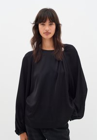 Blusa nera fluida con scollatura tondeggiante, maniche lunghe a palloncino e texture morbida. Presenta delicate pieghe vicino alle spalle.