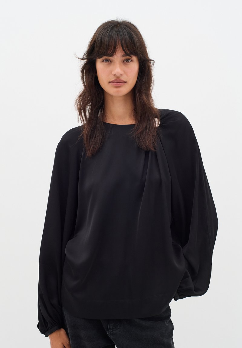 Blusa nera fluida con scollatura tondeggiante, maniche lunghe a palloncino e texture morbida. Presenta delicate pieghe vicino alle spalle.