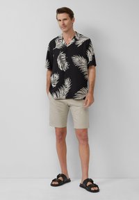 Chemise noire à manches courtes avec un motif de feuille de palmier blanche, associée à un short beige et des sandales noires. Tissu lisse, coupe décontractée.