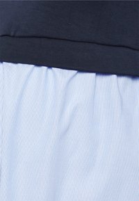 Haut bleu marine à encolure ronde, superposé à une jupe plissée bleu clair avec de fines rayures blanches verticales. Texture du textile visible.