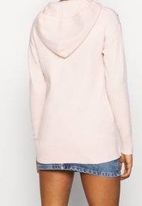 Person klädd i en ljusrosa huvtröja och en kort blå denimkjol, sedd bakifrån mot en enfärgad bakgrund.