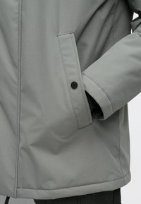 Šedá zateplená bunda s vysokým golierom, bočnými vreckami a čiernymi patentkami. Hladká, vodeodolná tkanina s moderným dizajnom.