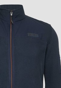 Marine blauwe zip-up jack met een textuur gebreide body en gladde stoffen schouderaccenten. Voorzien van een hoge kraag en contrasterende ritssluiting details.