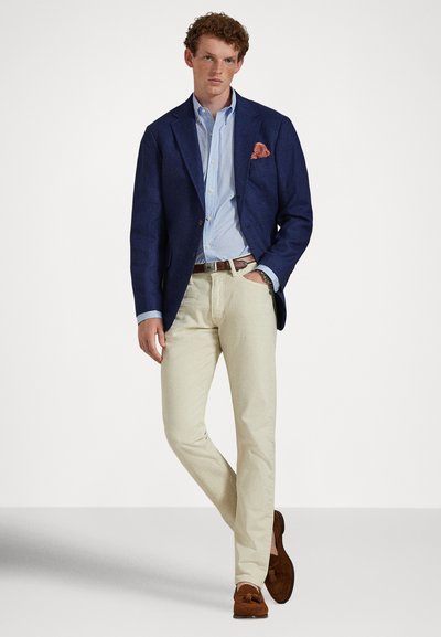 Young man wearing navy blazer with orange pocket square, light blue shirt, beige pants, brown belt, and brown loafers, standing with one hand in pocket.