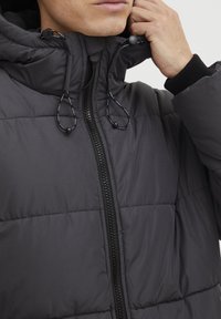 Veste rembourrée grise avec un col haut, fermeture éclair noire et cordons de serrage pour l'ajustement. Présente une texture matelassée et des poignets côtelés.