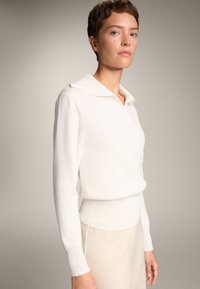 Pulover alb din tricot cu tiv ondulat și mâneci lungi, având un guler pliat. Textură moale, conceput pentru o croială lejeră.