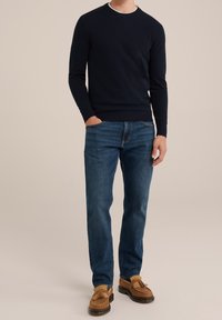 Man i mörknavy tröja, blå jeans och beige fransloafers med ena handen i fickan mot en neutral bakgrund.