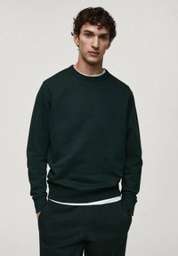Felpa verde scuro con colletto rotondo, polsini e vita a costine. Materiale in misto cotone, texture liscia, indossata sopra una t-shirt bianca.