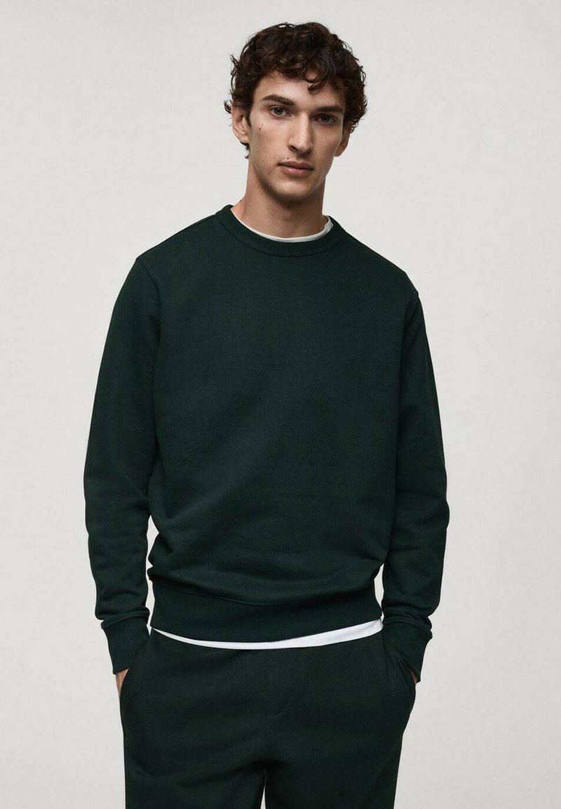 Felpa verde scuro con colletto rotondo, polsini e vita a costine. Materiale in misto cotone, texture liscia, indossata sopra una t-shirt bianca.