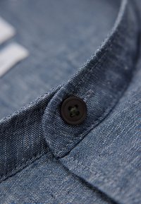 Närbild av kragen på en denimskjorta, med en svart knapp med en grön accent, texturerad blå tyg och noggranna sömnadsdetaljer.