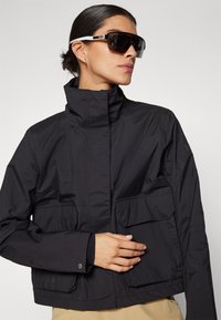 Chaqueta ligera negra con cuello alto, bolsillos frontales y una textura suave, combinada con grandes gafas de sol oscuras con acentos blancos.