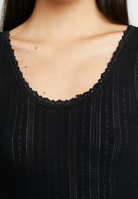 Top negro sin mangas con textura acanalada, que presenta un borde de encaje con forma de concha en el escote y patrones de rayas verticales en toda la prenda.