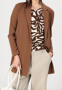 Blazer marrone sopra un top a fantasia crema e marrone, abbinato a pantaloni beige. La modella tiene una borsa bianca. Tessuto liscio, vestibilità su misura.