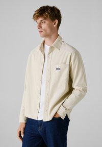 Beige corduroy shirt met een button-down ontwerp, borstzak en blauw "Red Bull Racing" logo. Gedeeltelijk gedragen over een wit t-shirt met donkere jeans.
