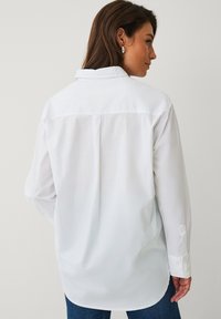 Chemise blanche boutonnée avec un col pointu, manches longues, et un dos droit. Tissu doux avec une texture lisse.