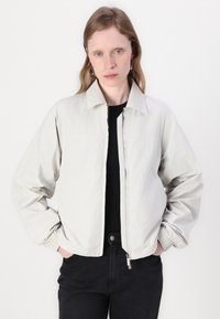 Hellgraue Bomberjacke mit Reißverschluss, umklappbarem Kragen, elastischen Bündchen und lässiger Passform. Kombiniert mit einem schwarzen Oberteil und dunklen Jeans.