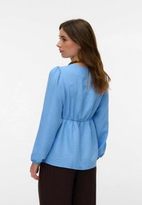 Blouse bleu clair en tissu texturé, col rond, taille froncée et petite découpe au dos. Manches longues avec poignets élastiques.