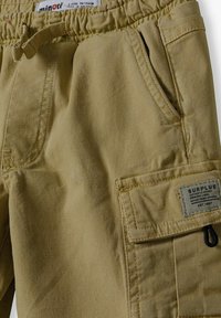 Shorts cargo beiges avec une ceinture élastique, plusieurs poches et un tissu texturé. Comprend une étiquette avec le logo "Surplus".