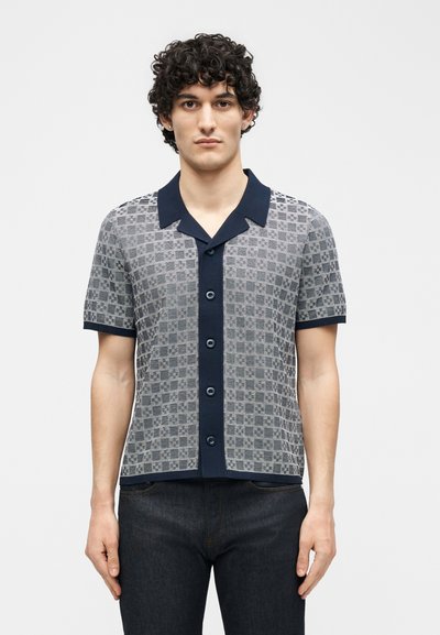 Jeune homme aux cheveux bouclés portant une chemise à manches courtes gris et bleu marine à boutons avec un motif géométrique et un jean foncé, debout face à l'avant.