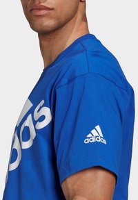 Modrý bavlněný tričko s velkým bílým logem "adidas", krátkými rukávy, kulatým výstřihem a malým bílým logem na levém rukávu.