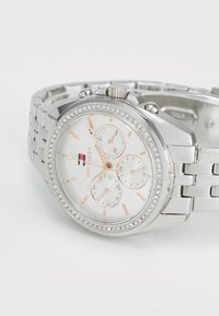 Montre en acier inoxydable argenté avec un cadran rond blanc, des aiguilles en or rose et trois sous-cadrans, soulignée par une lunette ornée de strass.