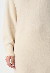 Mano che indossa un maglione a costine beige con polsino ripiegato, appoggiata accanto al torso su uno sfondo bianco.