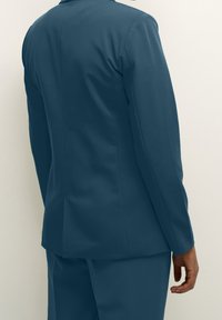 Turkusowa marynarka o dopasowanym kroju, gładkiej fakturze i wąskich klapach, z pojedynczym rozcięciem z tyłu i długimi rękawami.