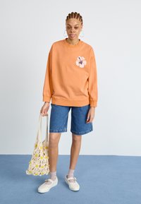 Sweatshirt pêche avec un design floral, short en denim, chaussures crème et sac en crochet avec des accents jaunes. Texture lisse et coupe décontractée.