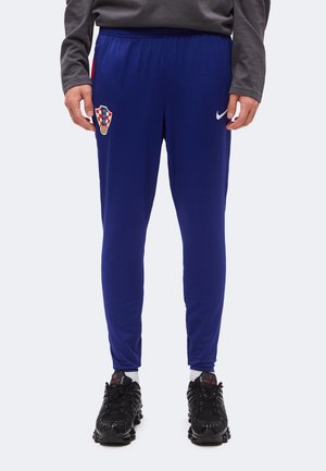 Personne portant un pantalon de sport bleu avec un emblème de football croate et un logo Nike blanc, des baskets noires et un t-shirt gris à manches longues.