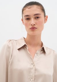 Blusa in satin beige con colletto classico, sottili pieghe sulle spalle e bottoni decorativi simili a cristalli. Texture liscia con una finitura elegante.