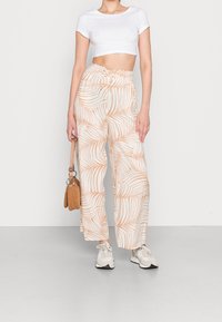 Lightweight, wide-leg trousers with a beige tropical leaf pattern. Paired with a white cropped top and sneakers. Accented by a small handbag.